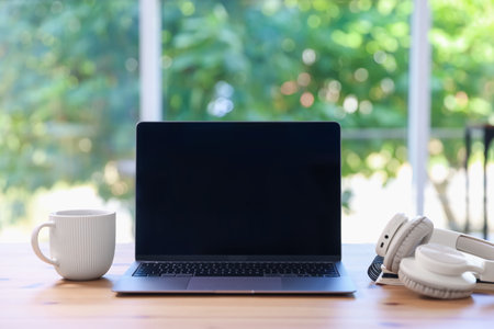 Home workspace. Laptop, cup of drink and headphones on wooden desk near window indoors
