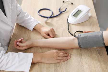 Doctor checking woman's heart rate and blood pressure via sphygmomanometer at wooden table indoors, closeupの写真素材