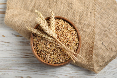 Wheat grains in bowl, spikes and burlap cloth on light wooden table, top viewの写真素材