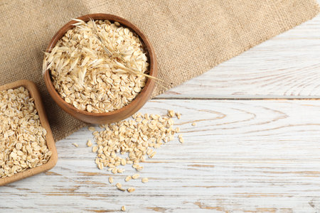 Oat flakes in bowl, spoon, spikes and burlap cloth on light wooden table, flat lay. Space for text