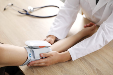 Doctor checking woman's heart rate and blood pressure via sphygmomanometer at wooden table indoors,...