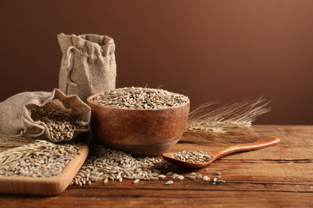 Rye grains in bowl, sack, spoon and spikes on wooden table against brown background, closeupの写真素材