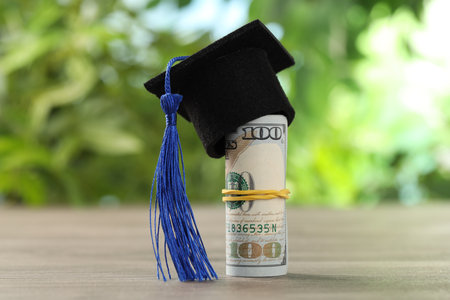 Scholarship. Rolled dollar banknotes with graduate hat on wooden table outdoors, closeupの写真素材