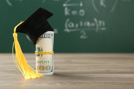 Scholarship. Rolled dollar banknotes with graduate hat on wooden table against green chalkboard, closeup. Space for textの写真素材