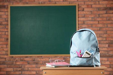 Light blue backpack with different stationery and glasses on desk in classroom. Space for textの写真素材