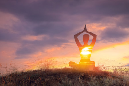 Practicing yoga in nature. Woman meditating in lotus position on hill. Double exposure with sunset skyの写真素材