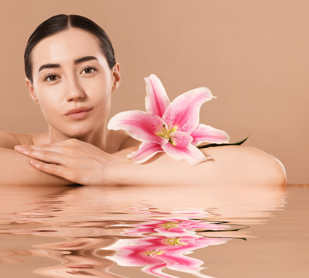 Beautiful woman with perfect skin and lily flower against pale brown background. Her reflection in rippled waterの写真素材
