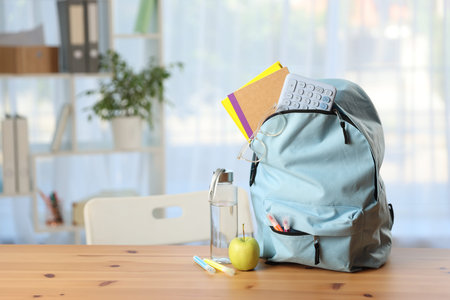 Backpack, different stationery, bottle of water and apple on desk in classroom. Space for text