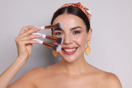 Beautiful woman with makeup brushes on light gray background