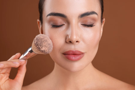 Beautiful woman applying makeup with brush on brown background, closeup