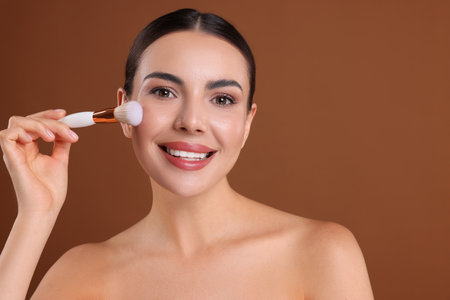 Beautiful woman applying makeup with brush on brown background, space for textの写真素材