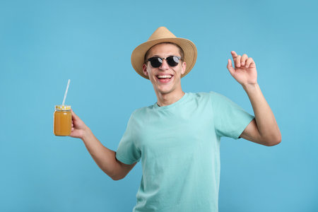 Happy man in sunglasses and hat with mason jar of orange juice on light blue background. Refreshing drinkの写真素材