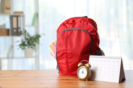 Red backpack with different stationery, calendar and alarm clock on desk in classroomの写真素材