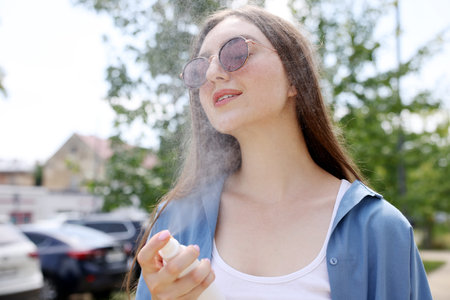 Smiling woman in sunglasses spraying thermal water onto her face outdoors