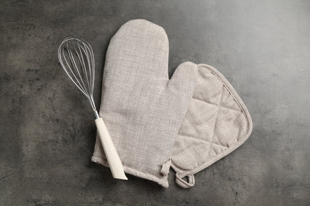 Grey oven glove, potholder and whisk on dark textured table, flat layの写真素材