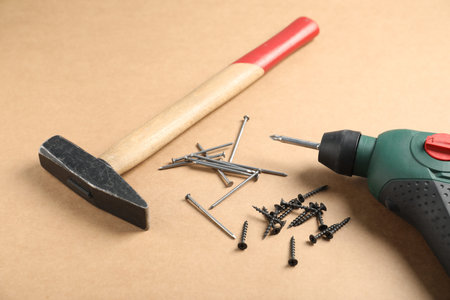 Metal nails, screws, hammer and electric screwdriver on cardboard surface, closeup