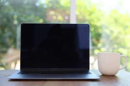 Open laptop and cup on wooden table against blurred green background, closeup. Mockup for design