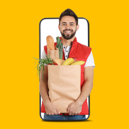 Delivery service worker giving online order out of mobile phone against orange background. Man with paper bag full of food productsの写真素材