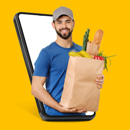 Delivery service worker giving online order out of mobile phone against orange background. Man with paper bag full of food productsの写真素材