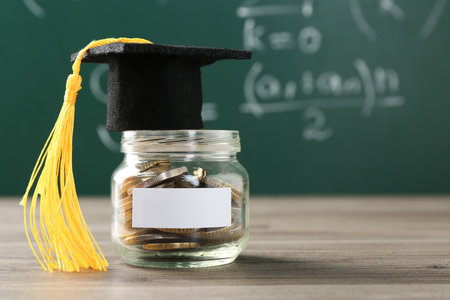 Scholarship. Jar with graduate hat and coins on wooden table against green chalkboard, closeup. Space for textの写真素材