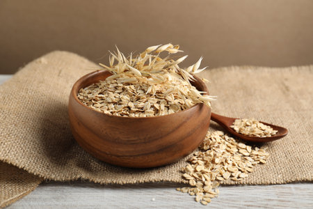 Oat flakes in bowl, spoon, spikes and burlap cloth on light wooden table against beige background,...