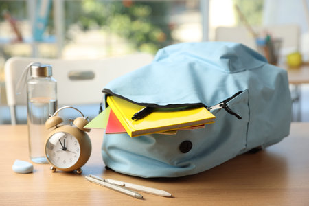 Backpack with different stationery, alarm clock and bottle of water on desk in classroom, closeup