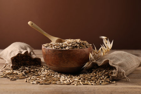 Oat grains, spikes, bowl and sacks on wooden table against brown background, closeupの写真素材