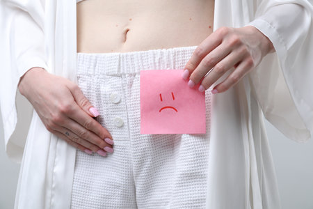 Woman holding sticky note with drawn sad face and suffering from cystitis symptoms on light gray background, closeupの写真素材