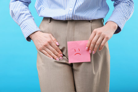 Woman holding sticky note with drawn sad face and suffering from cystitis symptoms on light blue background, closeupの写真素材