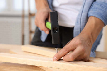 Man hammering nail into wooden plank indoors, closeup. Space for textの写真素材