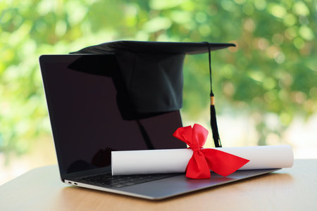 Graduation cap, laptop and diploma on wooden table against blurred green background, closeupの写真素材