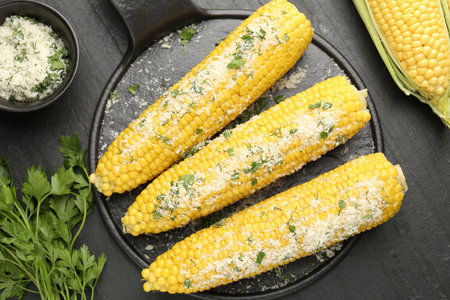 Tasty grilled corncobs with parsley and cheese on black table, flat lay