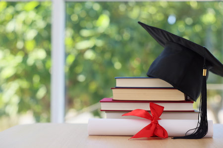Graduation cap, stack of books and diploma on wooden table against blurred green background, closeup. Space for textの写真素材