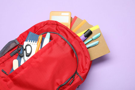 Red backpack with different stationery on violet background, top view