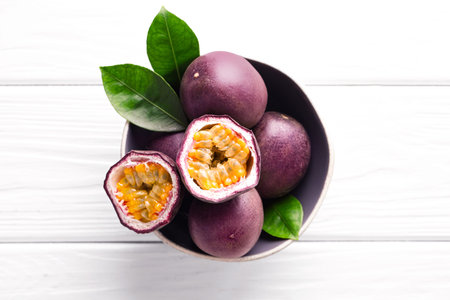 Fresh passion fruits and green leaves in bowl on white wooden table, top view