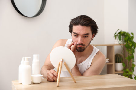 Young man with acne problem looking at mirror at table indoorsの写真素材