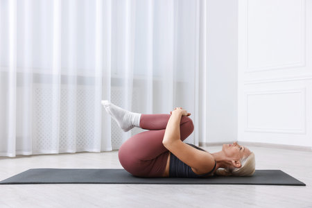 Mature woman in sportswear stretching on mat near window at home