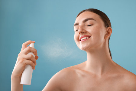 Smiling woman spraying thermal water onto her face on light blue backgroundの写真素材