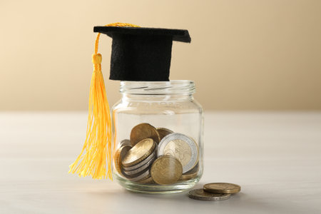 Scholarship. Jar with graduate hat and coins on white wooden table against beige background, closeupの写真素材