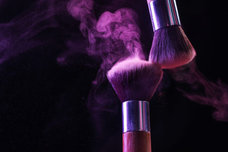 Makeup brushes with purple eye shadow in neon lights on black background, closeup. Space for text