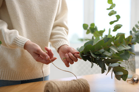 Florist with spool of thread making beautiful bouquet of eucalyptus branches at wooden table in...