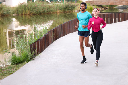 Sporty couple in headphones running near river outdoors. Space for text