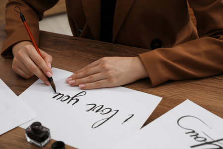 Calligraphy. Woman with brush writing phrase I Love You on sheet of paper at wooden table, closeupの写真素材