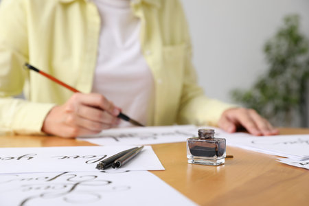Calligraphy. Woman with brush writing at wooden table indoors, focus on inkwell and fountain pens
