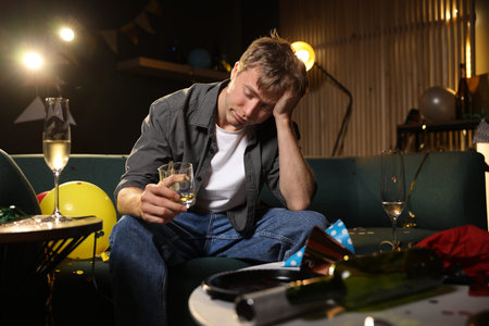Man with glass of alcohol suffering from hangover on sofa in messy room after party