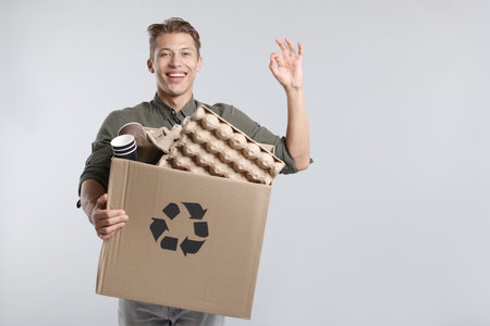 Recycling. Smiling man holding cardboard box with different paper waste and showing OK gesture on light background. Space for textの写真素材