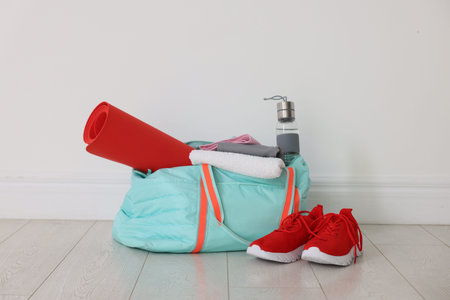 Stylish sports bag with sportswear, fitness mat, towel, water bottle and sneakers on floor near...