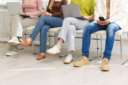 People with different devices waiting in queue on chairs indoors, closeup