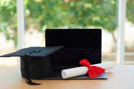 Graduation cap, laptop and diploma on wooden table against blurred green background, closeupの写真素材