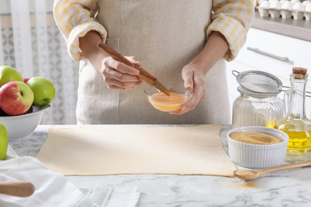 Making delicious apple strudel. Woman spreading egg yolk onto dough at white marble table in...